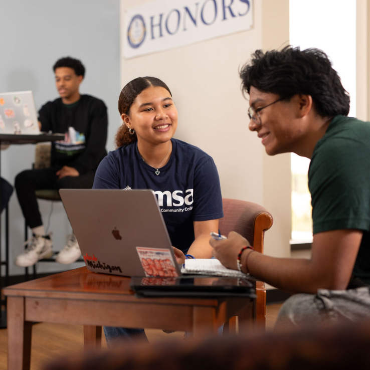 A group of three students collaborate on schoolwork in a study space, with a world map on the wall and a sign reading "HONORS" above.